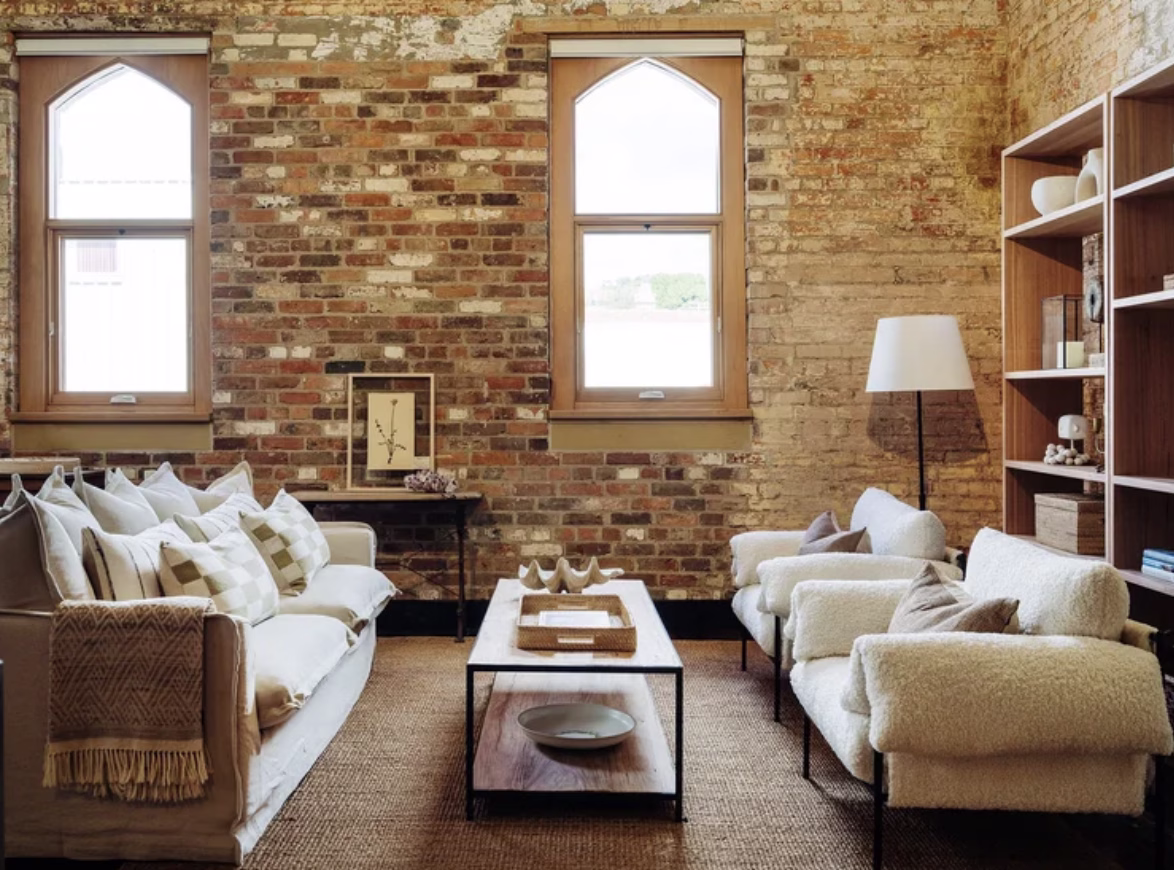 The photograph of the interior of one of The Rox's apartments displays a seating area with two plush chairs opposite a couch with a coffee table in between. Exposed brick, lots of neutrals, and light-filled windows complete the shot.